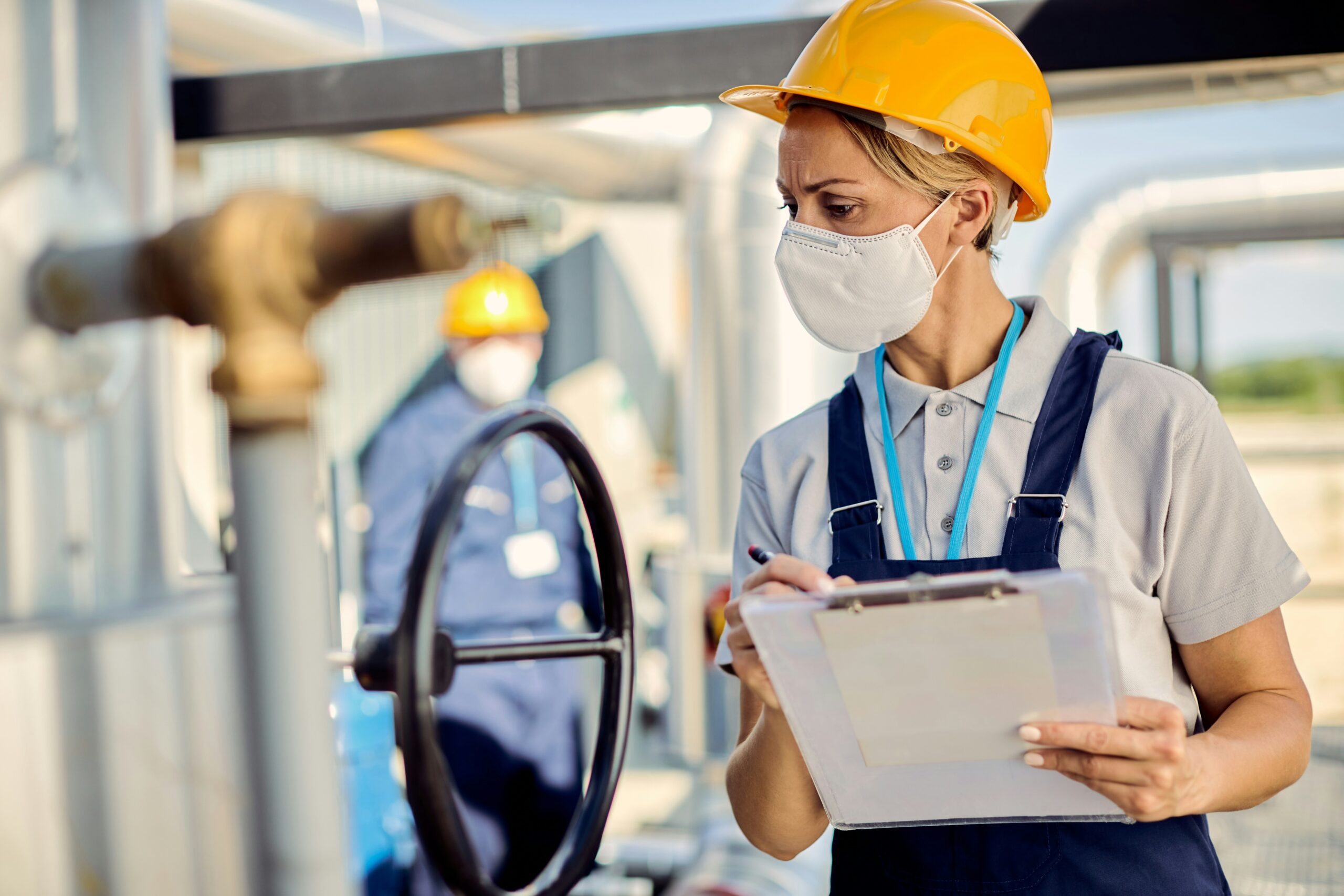 Woman wearing white mask and yellow hard hat holding clipboard